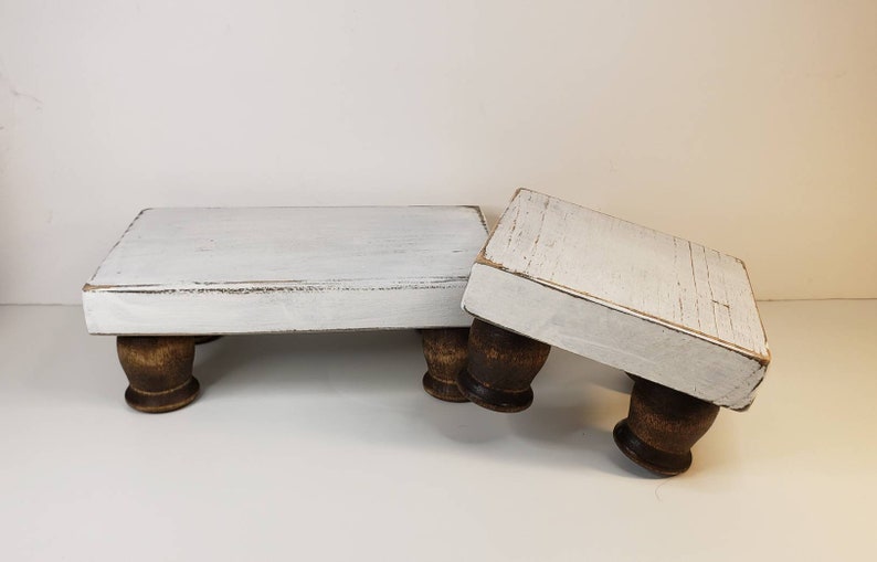 May include: Two white wooden tiered stands with distressed paint and brown wooden legs. The stands are stacked on top of each other.