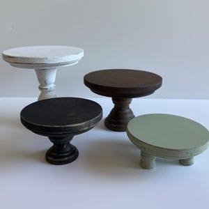Puede incluir: Un conjunto de cuatro peque&ntilde;os soportes de madera con tapas circulares y bases de pedestal o patas cortas. El set incluye un soporte blanco desgastado, uno marr&oacute;n oscuro, uno negro y un soporte bajo de color verde salvia.