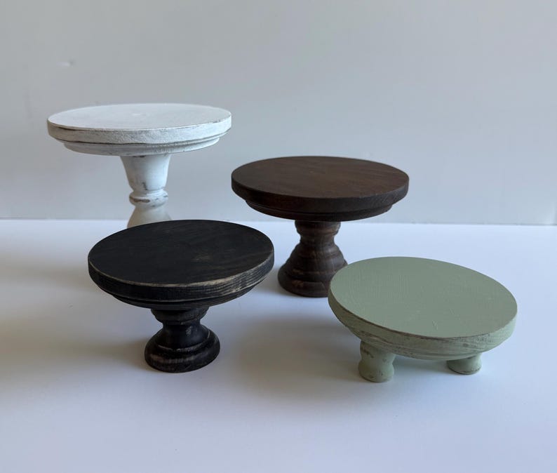 Puede incluir: Un conjunto de cuatro peque&ntilde;os soportes de madera con tapas circulares y bases de pedestal o patas cortas. El set incluye un soporte blanco desgastado, uno marr&oacute;n oscuro, uno negro y un soporte bajo de color verde salvia.