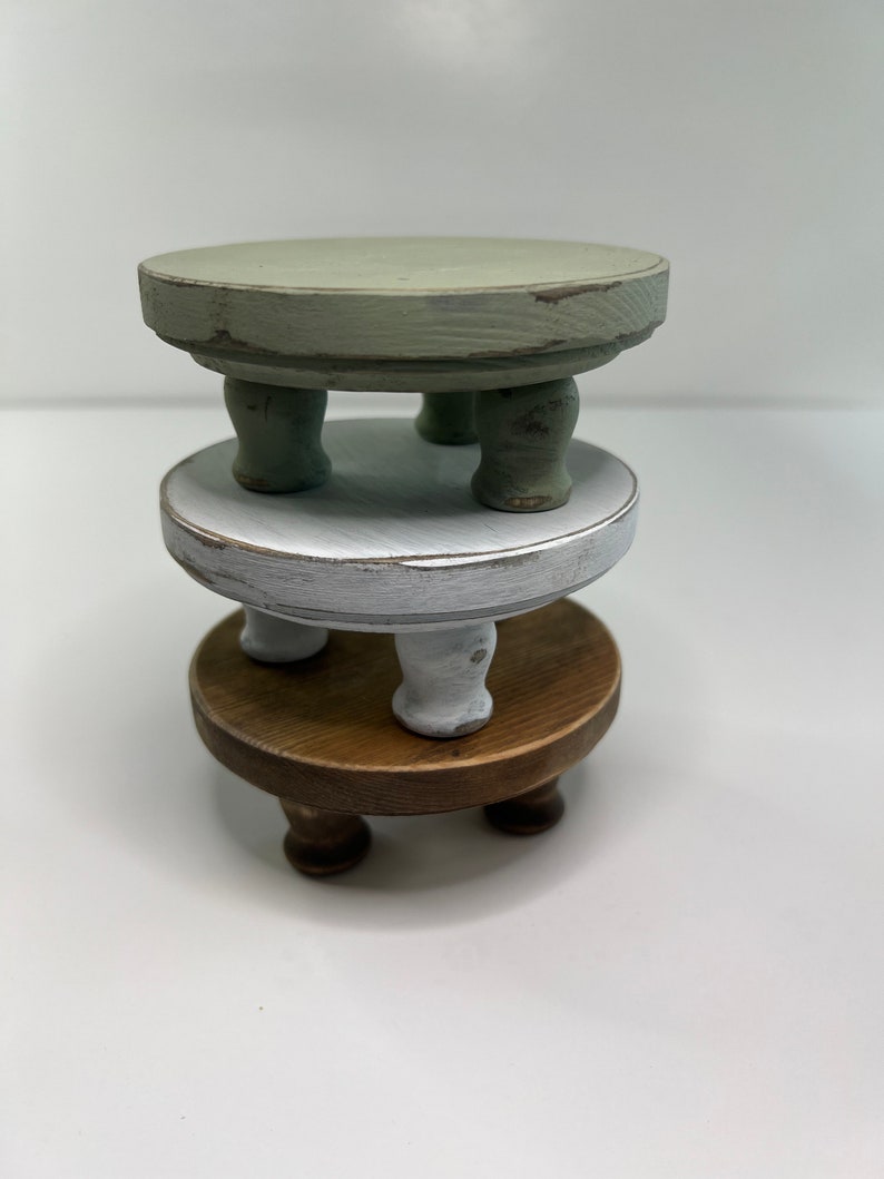 Puede incluir: Una pila de tres elevadores redondos de madera con patas cortas torneadas. El elevador superior est&aacute; pintado de verde salvia, el del medio es blanco con un acabado desgastado y el inferior presenta un acabado de veta de madera natural oscura.