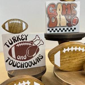 May include: A tiered wooden stand with four brown footballs, two with white laces. The top tier has a white sign with the text "GAME DAY" in brown letters. The middle tier has a white sign with the text "TURKEY and TOUCHDOWNS" in brown letters, with a brown football and a turkey illustration. The bottom tier has a white sign with brown text and a leopard print background.