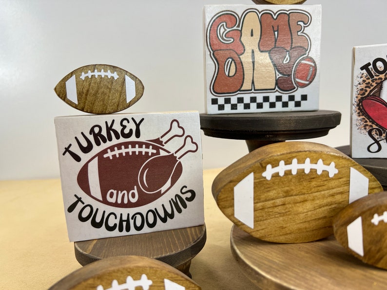 May include: A tiered wooden stand with four brown footballs, two with white laces. The top tier has a white sign with the text "GAME DAY" in brown letters. The middle tier has a white sign with the text "TURKEY and TOUCHDOWNS" in brown letters, with a brown football and a turkey illustration. The bottom tier has a white sign with brown text and a leopard print background.