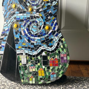 K&ouml;nnte beinhalten: Eine Mosaikgitarre mit einem Design, das von Van Goghs "Sternennacht" inspiriert wurde. Das Kunstwerk verwendet blaue, gelbe und gr&uuml;ne Fliesen, um einen wirbelnden Nachthimmel, Sterne und ein Dorf darzustellen. Der Korpus der Gitarre ist schwarz.