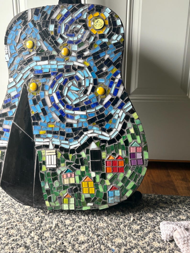 K&ouml;nnte beinhalten: Eine Mosaikgitarre mit einem Design, das von Van Goghs "Sternennacht" inspiriert wurde. Das Kunstwerk verwendet blaue, gelbe und gr&uuml;ne Fliesen, um einen wirbelnden Nachthimmel, Sterne und ein Dorf darzustellen. Der Korpus der Gitarre ist schwarz.