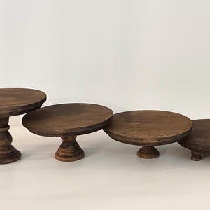 Puede incluir: Un conjunto de cuatro soportes circulares de madera marr&oacute;n oscuro con alturas y estilos de base variados. Los soportes cuentan con superficies planas y lisas, dispuestos en fila, mostrando dise&ntilde;os de pedestal desde altos hasta bajos.