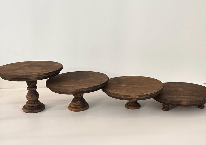 Puede incluir: Un conjunto de cuatro soportes circulares de madera marr&oacute;n oscuro con alturas y estilos de base variados. Los soportes cuentan con superficies planas y lisas, dispuestos en fila, mostrando dise&ntilde;os de pedestal desde altos hasta bajos.