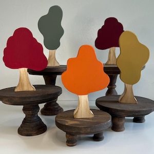 May include: Five wooden tree figurines with painted leaves in various colors, including red, orange, yellow, and green, are displayed on individual wooden pedestal stands.