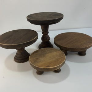 Puede incluir: Un conjunto de cuatro soportes de madera marr&oacute;n oscuro con tapas circulares y pies de pedestal o cortos. La colecci&oacute;n incluye un soporte alto, un soporte mediano y dos soportes bajos, todos con un acabado de veta de madera visible.