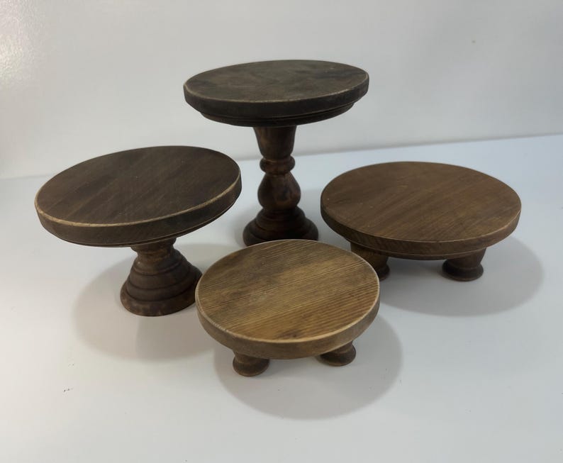 Puede incluir: Un conjunto de cuatro soportes de madera marr&oacute;n oscuro con tapas circulares y pies de pedestal o cortos. La colecci&oacute;n incluye un soporte alto, un soporte mediano y dos soportes bajos, todos con un acabado de veta de madera visible.