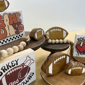 May include: A tiered wooden stand with three levels, each decorated with a different football-themed sign and wooden football ornaments. The top level features a sign that says "Game Day", the middle level says "Turkey and Touchdowns", and the bottom level says "Touchdown Season".