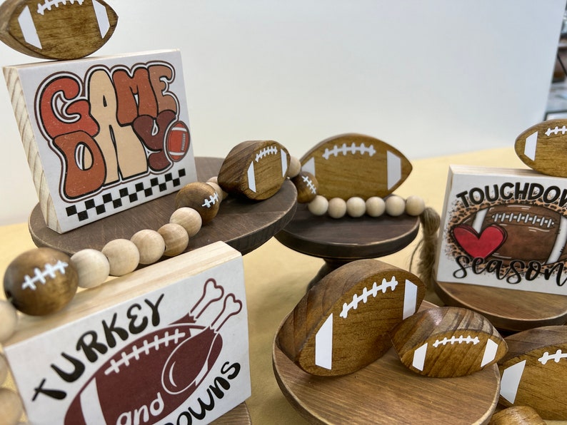 May include: A tiered wooden stand with three levels, each decorated with a different football-themed sign and wooden football ornaments. The top level features a sign that says "Game Day", the middle level says "Turkey and Touchdowns", and the bottom level says "Touchdown Season".