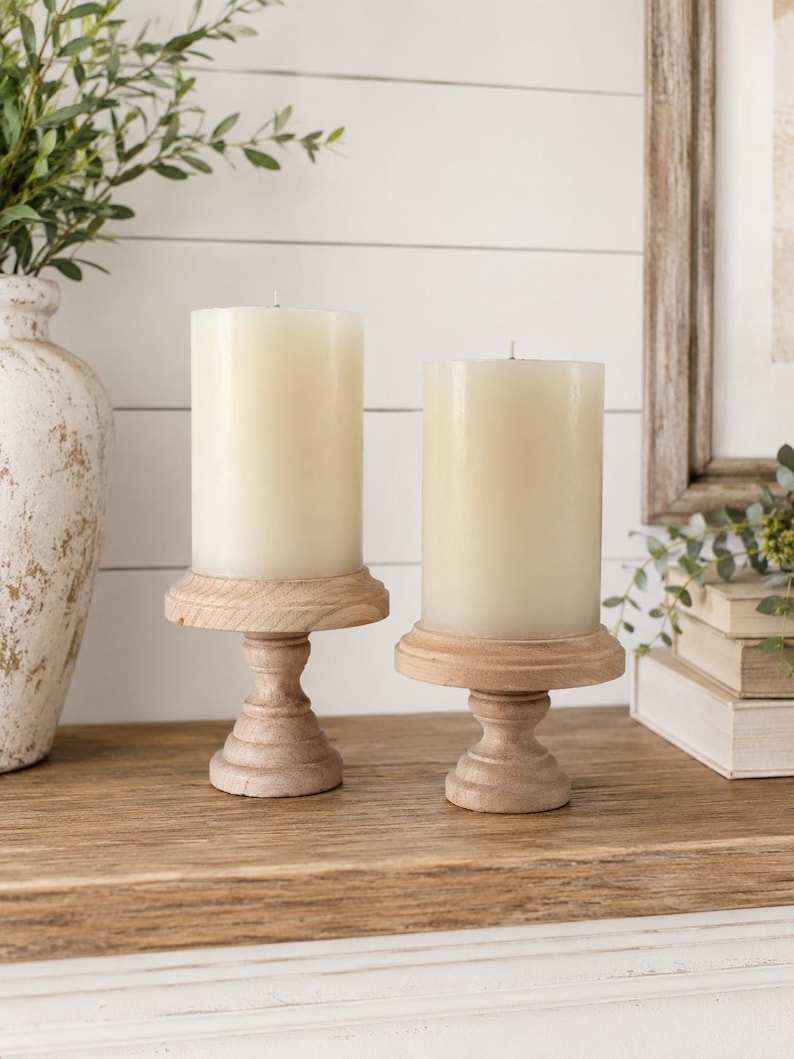 May include: Two ivory-colored pillar candles sit on light brown wooden candle holders. The candle holders have a pedestal base and a round top. The candles are unscented and cylindrical, creating a warm, inviting ambiance.