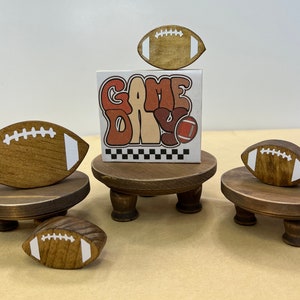May include: Three wooden stands with footballs on top. The stands are painted brown and have three legs. The footballs are painted brown with white stitching. The middle stand has a sign that says "Game Day" in a retro font.