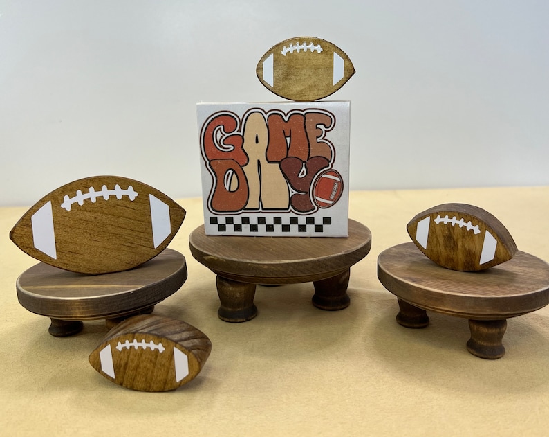 May include: Three wooden stands with footballs on top. The stands are painted brown and have three legs. The footballs are painted brown with white stitching. The middle stand has a sign that says "Game Day" in a retro font.