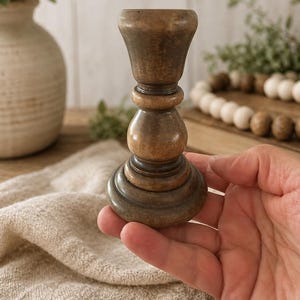 Peut inclure: Un bougeoir en bois tourn&eacute; avec une finition marron fonc&eacute;. Le bougeoir a une base large et &eacute;tag&eacute;e, une section m&eacute;diane arrondie et un dessus &eacute;vas&eacute;. Le support est tenu dans une main, sugg&eacute;rant sa taille et sa portabilit&eacute;. Le style est rustique et vintage.