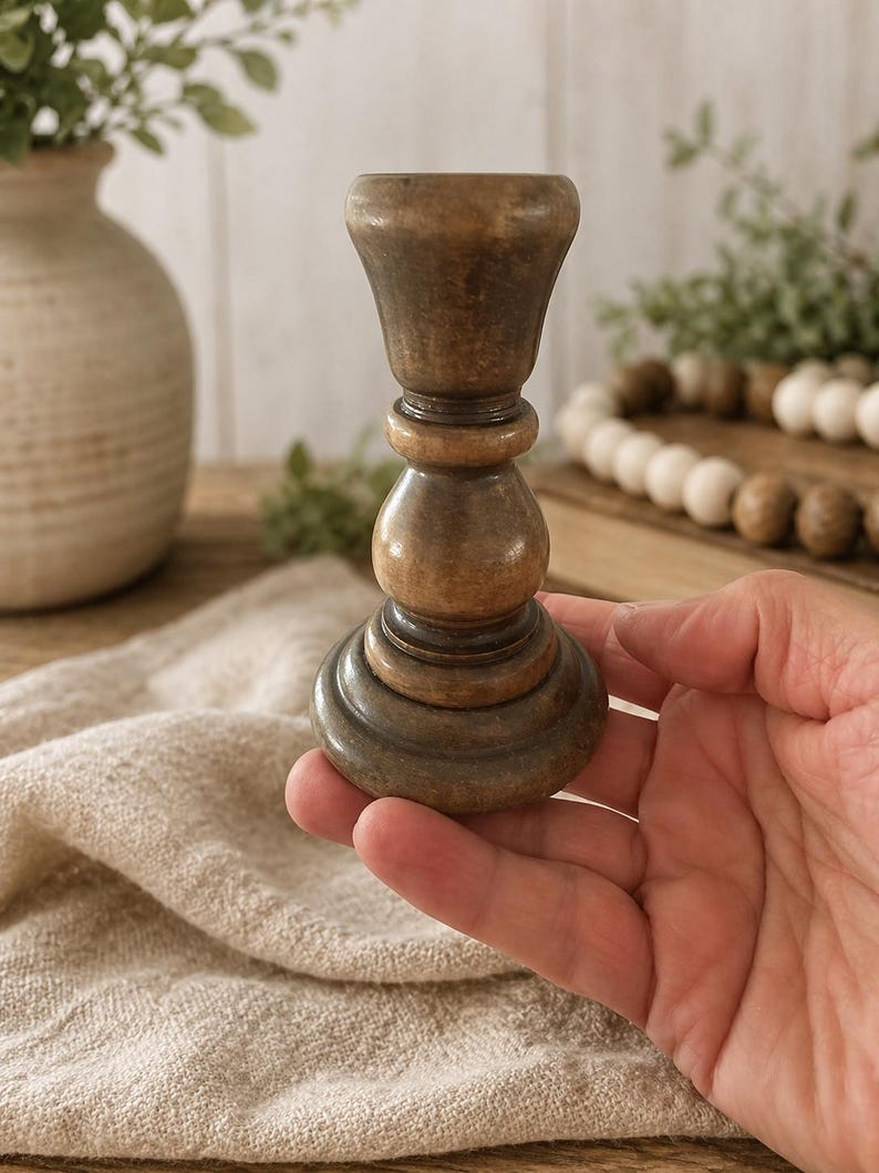 Peut inclure: Un bougeoir en bois tourn&eacute; avec une finition marron fonc&eacute;. Le bougeoir a une base large et &eacute;tag&eacute;e, une section m&eacute;diane arrondie et un dessus &eacute;vas&eacute;. Le support est tenu dans une main, sugg&eacute;rant sa taille et sa portabilit&eacute;. Le style est rustique et vintage.