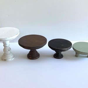 Puede incluir: Un conjunto de cuatro elevadores de madera de diferentes alturas y colores. El primero es un pedestal blanco alto, el segundo es un soporte marr&oacute;n oscuro mediano, el tercero es un soporte marr&oacute;n oscuro m&aacute;s corto y el cuarto es una base redonda baja de color verde salvia.