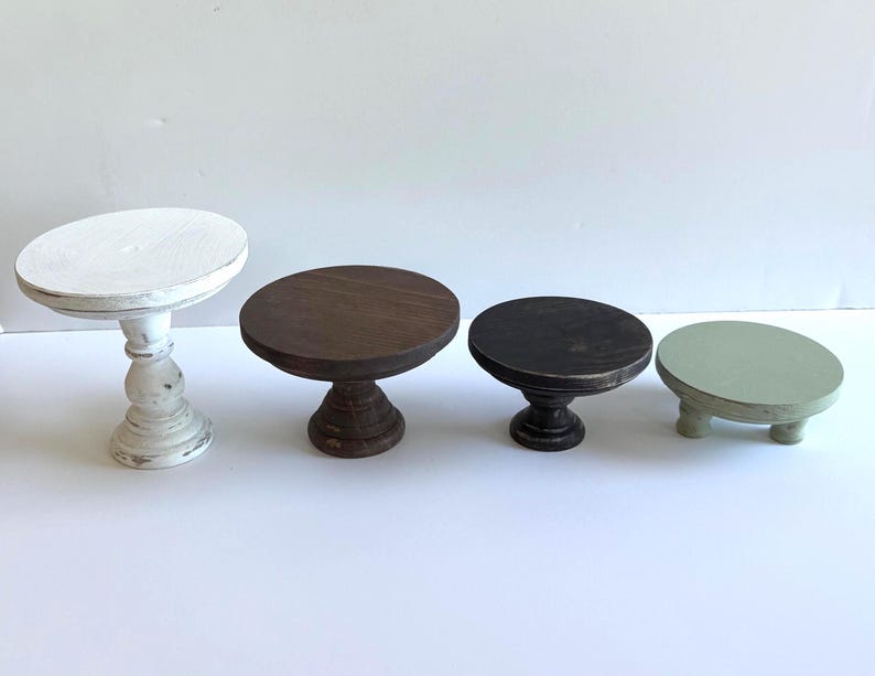 Puede incluir: Un conjunto de cuatro elevadores de madera de diferentes alturas y colores. El primero es un pedestal blanco alto, el segundo es un soporte marr&oacute;n oscuro mediano, el tercero es un soporte marr&oacute;n oscuro m&aacute;s corto y el cuarto es una base redonda baja de color verde salvia.