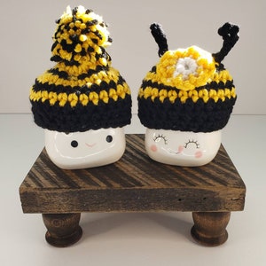 May include: Two white ceramic mugs with smiling faces wearing crocheted bee hats. The hats are black and yellow with a white pom-pom on one hat and black antennae on the other. The mugs are sitting on a wooden stand with two legs.