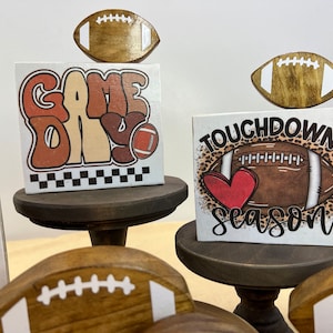 May include: Three wooden signs with football themes. The top sign says "Game Day" with a football graphic. The middle sign says "Touchdown Season" with a football and heart graphic. The bottom sign has a turkey leg graphic and the word "Lums" on it.