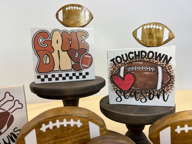 May include: Three wooden signs with football themes. The top sign says "Game Day" with a football graphic. The middle sign says "Touchdown Season" with a football and heart graphic. The bottom sign has a turkey leg graphic and the word "Lums" on it.