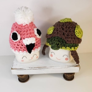 May include: Two white ceramic mugs with painted faces wearing crocheted hats. The mug on the left has a pink and white flamingo hat with black eyes and beak. The mug on the right has a brown and green turtle hat with green accents.