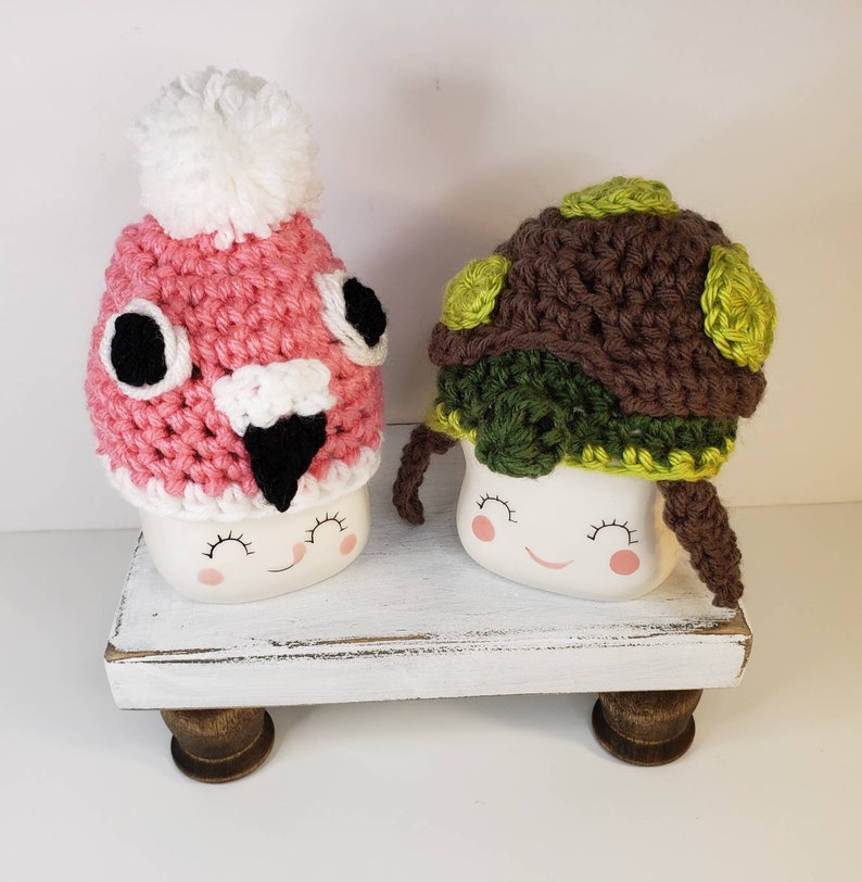 May include: Two white ceramic mugs with painted faces wearing crocheted hats. The mug on the left has a pink and white flamingo hat with black eyes and beak. The mug on the right has a brown and green turtle hat with green accents.