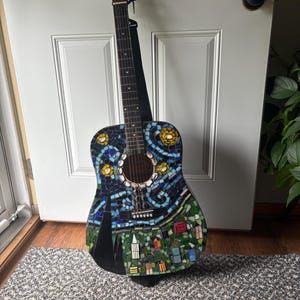 K&ouml;nnte beinhalten: Akustikgitarre mit Mosaikdesign. Der Korpus weist ein farbenfrohes Mosaikmuster mit blauen, gr&uuml;nen, gelben und wei&szlig;en Fliesen auf, das eine Sternennachtszene darstellt. Die Gitarre hat einen schwarzen Hals und einen schwarzen Gurt.