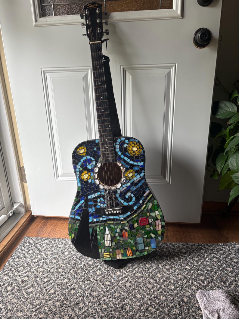 K&ouml;nnte beinhalten: Akustikgitarre mit Mosaikdesign. Der Korpus weist ein farbenfrohes Mosaikmuster mit blauen, gr&uuml;nen, gelben und wei&szlig;en Fliesen auf, das eine Sternennachtszene darstellt. Die Gitarre hat einen schwarzen Hals und einen schwarzen Gurt.