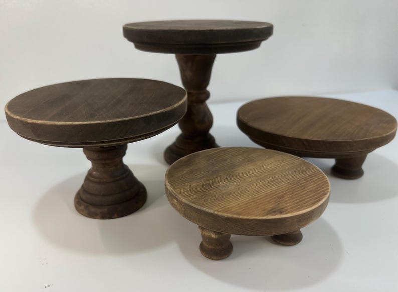 Puede incluir: Un conjunto de cuatro soportes de madera de color marr&oacute;n oscuro con superficies circulares y bases de pedestal de alturas variables. La colecci&oacute;n incluye dos soportes altos con columnas centrales y dos soportes m&aacute;s bajos con pies peque&ntilde;os.