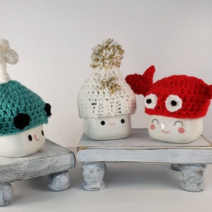 May include: Three white ceramic mugs with cute faces are wearing crocheted hats. The hats are a green narwhal, a white pom-pom beanie, and a red crab.