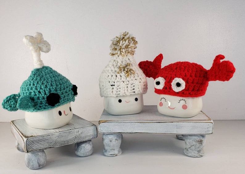 May include: Three white ceramic mugs with cute faces are wearing crocheted hats. The hats are a green narwhal, a white pom-pom beanie, and a red crab.