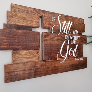May include: Wooden wall art with a cross shape made of multiple pieces of wood. The words "Be Still and Know That I Am God" are written in white cursive font with the verse "Psalm 46:10" below.