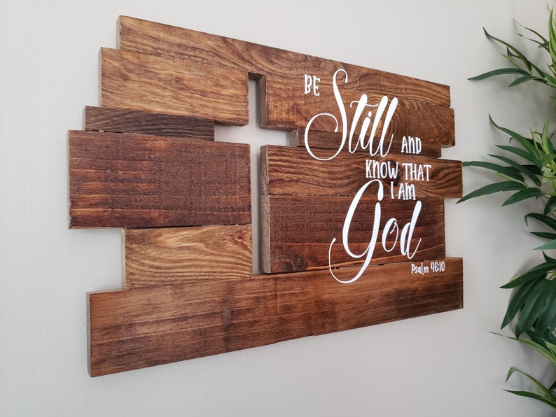 May include: Wooden wall art with a cross shape made of multiple pieces of wood. The words "Be Still and Know That I Am God" are written in white cursive font with the verse "Psalm 46:10" below.