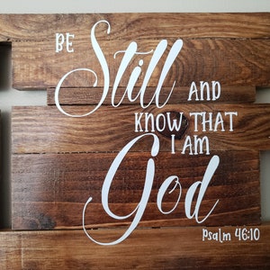 May include: Wooden sign with the words "Be still and know that I am God" in white lettering. The sign is made of multiple pieces of wood and has a rustic look. The verse is from Psalm 46:10.
