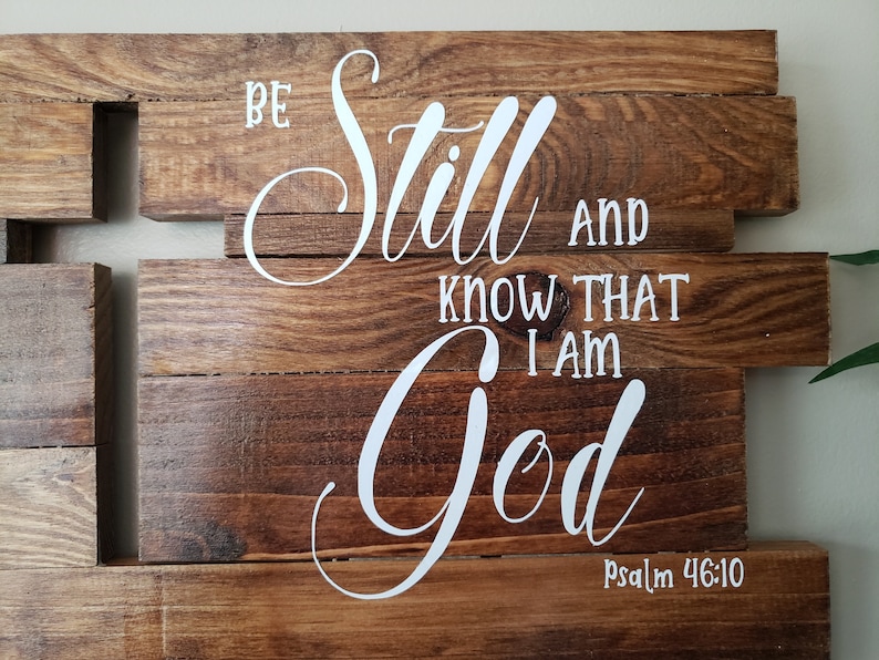 May include: Wooden sign with the words "Be still and know that I am God" in white lettering. The sign is made of multiple pieces of wood and has a rustic look. The verse is from Psalm 46:10.