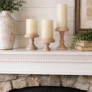 May include: Three ivory pillar candles on wooden pedestals, arranged on a white mantel. A decorative vase with greenery is on the left, and a framed picture and books are on the right. The fireplace below has a stone facade.