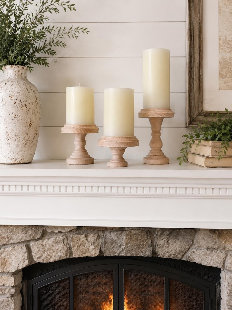 May include: Three ivory pillar candles on wooden pedestals, arranged on a white mantel. A decorative vase with greenery is on the left, and a framed picture and books are on the right. The fireplace below has a stone facade.