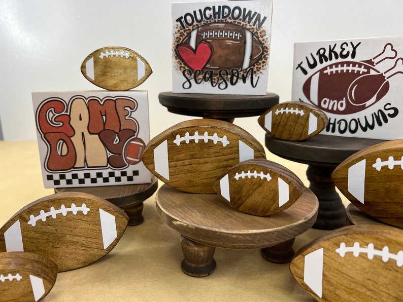 May include: A collection of wooden football-themed decorations, including signs that say "Game Day", "Touchdown Season", and "Turkey and Hoot", with a brown and white football on each sign.