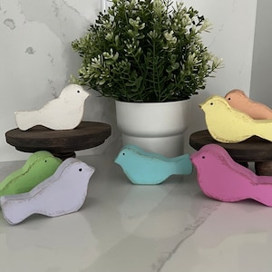 May include: A set of seven wooden bird figurines painted in pastel colors, including white, green, light blue, lavender, yellow, and orange. The birds are arranged on small wooden stands.