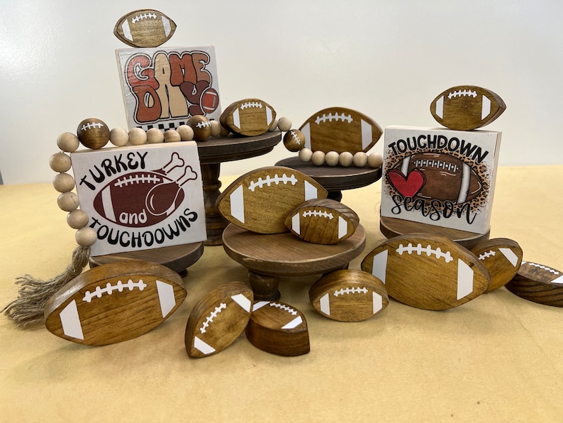 May include: Wooden signs with football and turkey themes, decorated with brown and white footballs. The signs say "Game Day", "Turkey and Touchdowns", and "Touchdown Season".