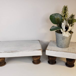 May include: Two white wooden tiered stands with distressed paint and brown wooden legs. The top tier has a small metal pot with a white flower and green foliage.