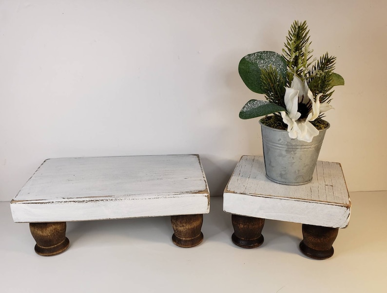 May include: Two white wooden tiered stands with distressed paint and brown wooden legs. The top tier has a small metal pot with a white flower and green foliage.