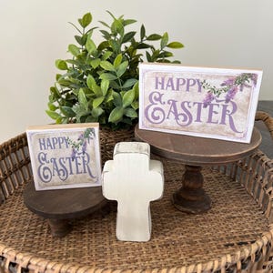 May include: Two wooden signs with the text "Happy Easter" in purple lettering with a floral design. A white wooden cross is in the center of the image. The signs and cross are on two small wooden stands.