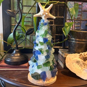 May include: A decorative tree made of sea glass in shades of blue, green, and white, topped with a white starfish. The tree sits on a round wooden base, creating a coastal or nautical theme.