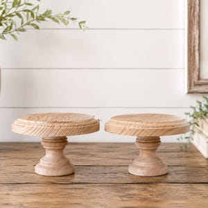 May include: Two small, round, wooden cake stands with a natural finish. Each stand has a pedestal base and a slightly raised top. The stands are displayed on a wooden surface, with a vase and greenery in the background.