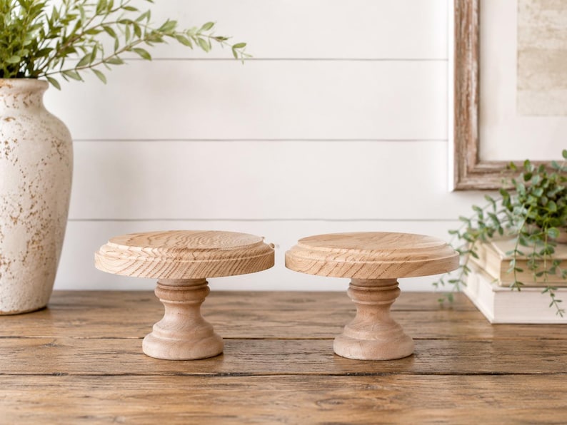 May include: Two small, round, wooden cake stands with a natural finish. Each stand has a pedestal base and a slightly raised top. The stands are displayed on a wooden surface, with a vase and greenery in the background.