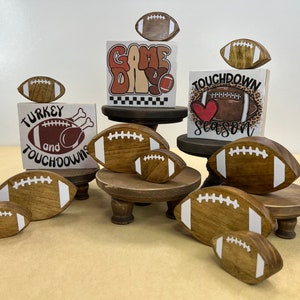 May include: A set of five wooden football-shaped ornaments with white stitching and text. The ornaments are displayed on three tiered wooden stands. The text on the ornaments reads "Game Day", "Turkey and Touchdowns", "Touchdown Season", and "Game Day".