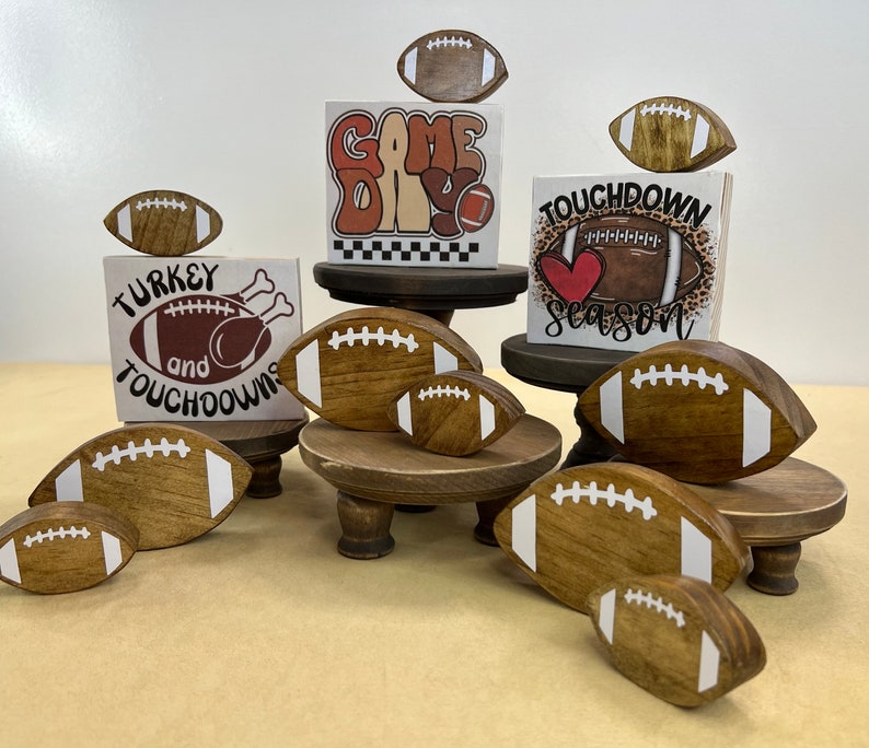 May include: A set of five wooden football-shaped ornaments with white stitching and text. The ornaments are displayed on three tiered wooden stands. The text on the ornaments reads "Game Day", "Turkey and Touchdowns", "Touchdown Season", and "Game Day".