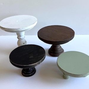 Puede incluir: Cuatro soportes circulares de madera con pedestal de diferentes alturas y colores. Un soporte es blanco con acabado desgastado, uno es marr&oacute;n oscuro, uno es negro y otro es de color verde salvia suave. Cada soporte tiene una base redonda.